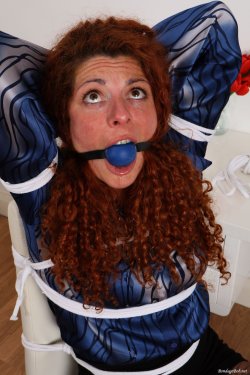 Curly haired red head Lola ball gagged and tied up in a blue silk blouse with her hands behind her head